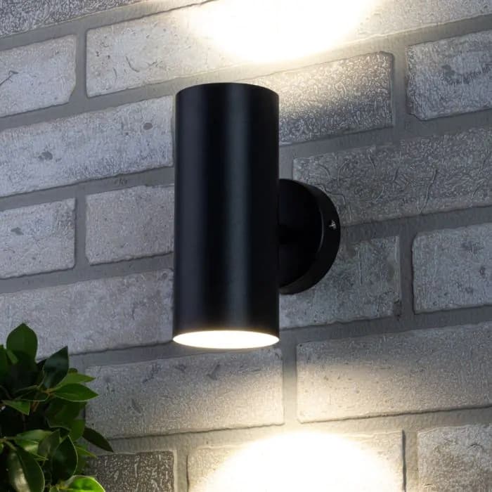 Black outdoor wall light fixture on a brick wall with a plant in the foreground.